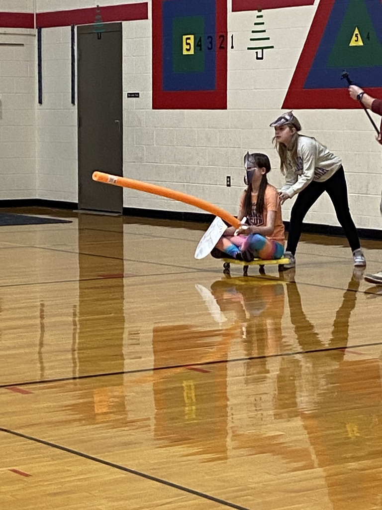 Students are sitting on a yellow board with wheels, holding an orange noodle and a shield wearing a mask.  Another student has her hands on the student who is sitting with the nooldle ready to push and pretend joust