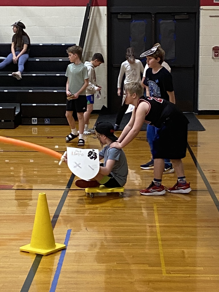 one student sitting on a yellow board with wheels, holding an orange pool noodle with a handle on it like a sword, wearing a mask and holding a shield.  The other student is behind him with his hands on his shoulders ready to push him.  Other students in the background are standing cheering on.  They are in the gym pretend jousting. 