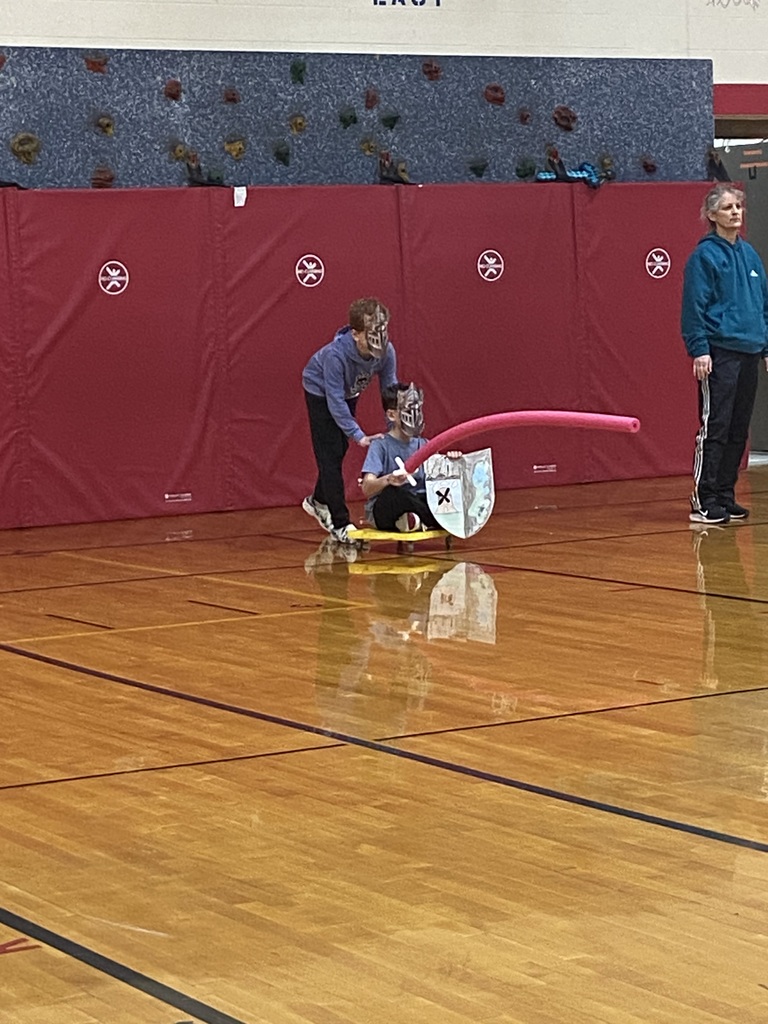 Two student in the gym.  One is sitting on a yellow board with wheels on it, holding a red pool noodle with a white handle, wearing a mask and holding a shield.  The other student is standing behind him wearing a mask and his hands are on the other students shoulder ready to push him.  The teacher is standing off to the side.  