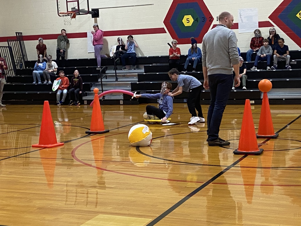 A student sitting on a yellow board with wheels holding a red pool noodle and a mask, a student behind him wearing a mask with his hands on the other students shoulders.  Teacher is standing watching the joustin tournament.  Orange cones have ball on top of them and students are sitting in the bleachers cheering them on. 