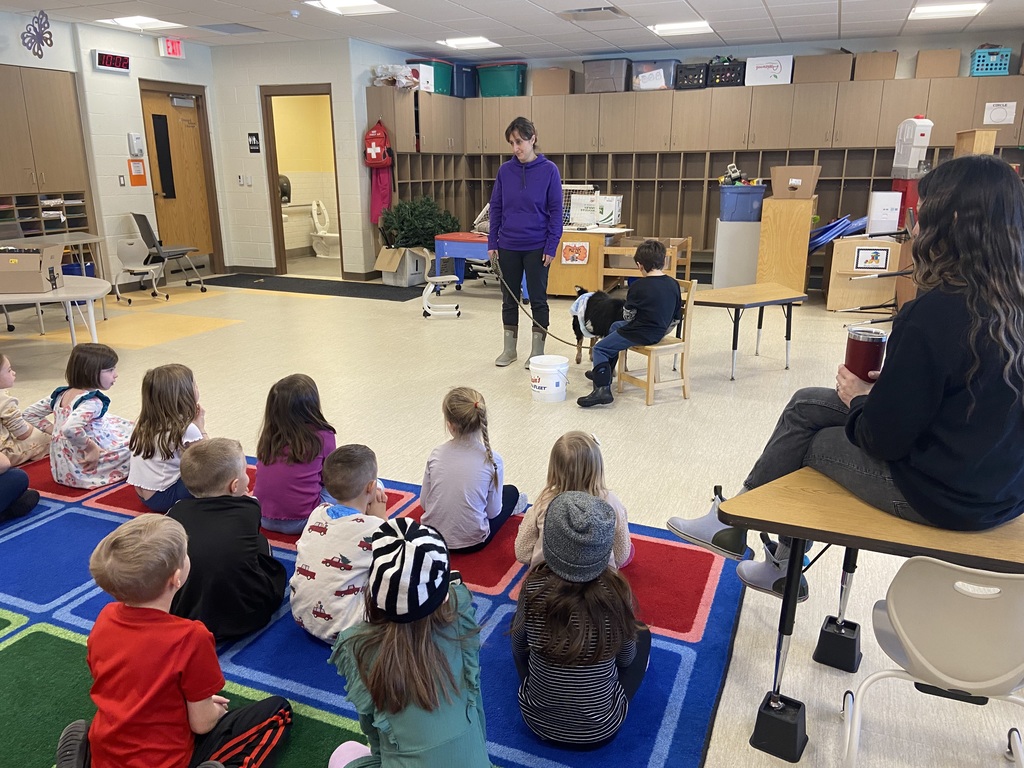 A teacher wearing a purple hoody holding a dwarf goat on a leash talking to students who are sitting at a blue, green and red rug listening to the teacher talk about the goat.  Another student is sitting on a chair nest to the goat with his back faced to the camera.  You can see the teacher sitting on a desk watching the presentation holding her maroon coffee mug. 
