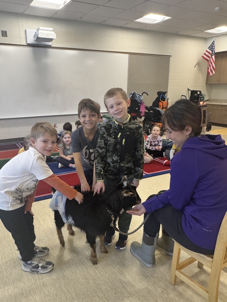 A teacher sitting on a chair wearing a purple sweatshirt, black pants, brown boots. while theree students are petting a dwarf goat on a leash.  The students are looking at the camera smiling.  The student on the left is wearing a white shirt with a graphic on it, with orange sleeves, black paints and running shoes.  The student in the middles is wearing a ble 6,7 shirt and the student on the right is wearing a green camo shirt with neon zipper pulls, black pants and black running shoes.  The students in the background are sitting on a square rug that has blue, red, green and purple squares. 