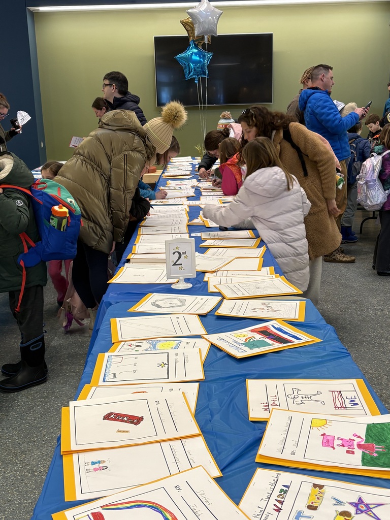 A row of tables with pieces of student writing lining both sides of the tables. Students and adults are leaning over to writing comments and read the writing.