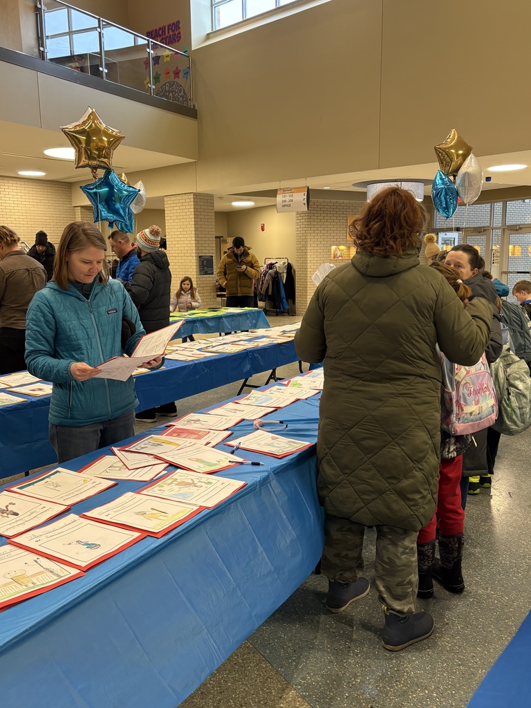 Part of two rows of tables with student writing on top of the tables. One adult is holding one of the writing pieces to read it. Other adults are standing on the sides of the tables to read student writing.