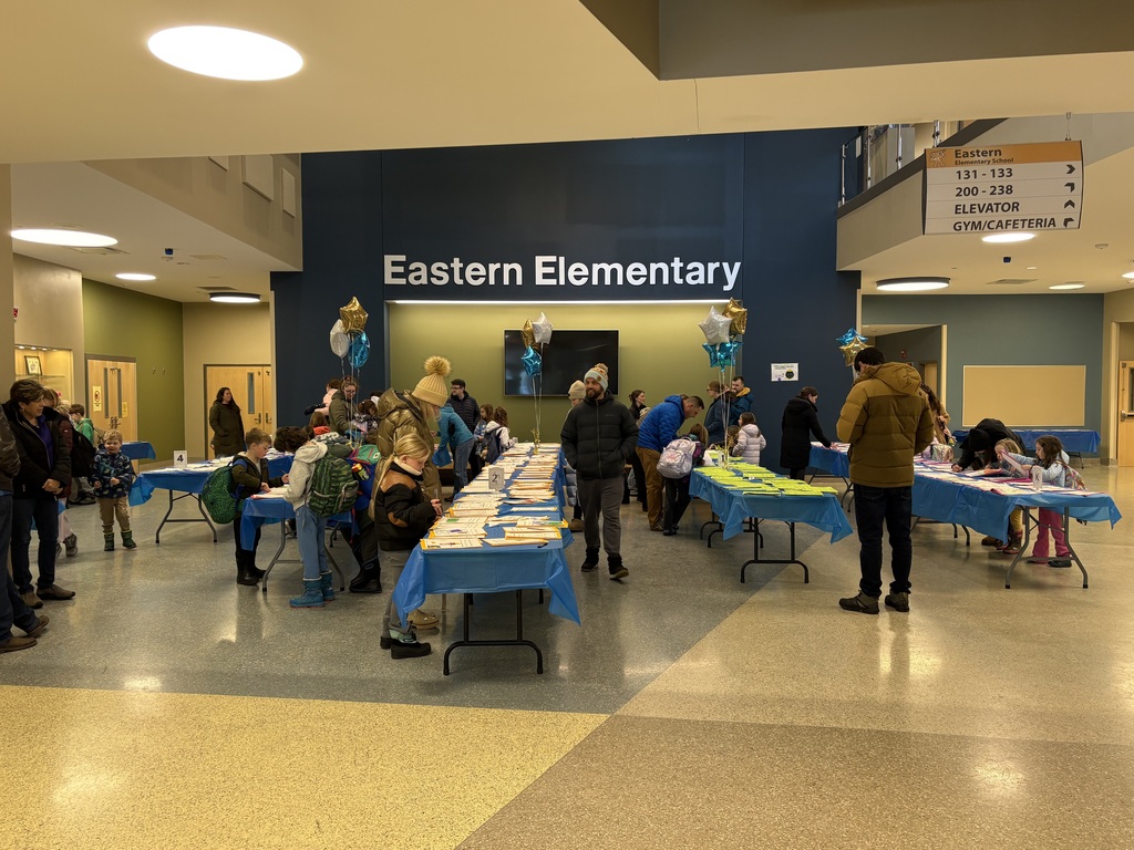 The Eastern Elementary lobby with four rows of long tables with student writing on the tables and students and adults on either side of the tables reading the writing and writing comments.