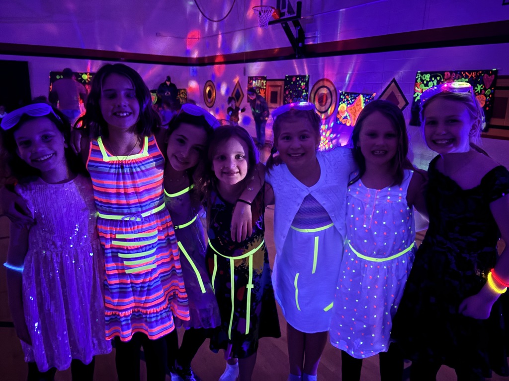 7 girls wearing glowing  fluorescent tape on their dresses with arms around each other in a gym with disco lights shining on the walls