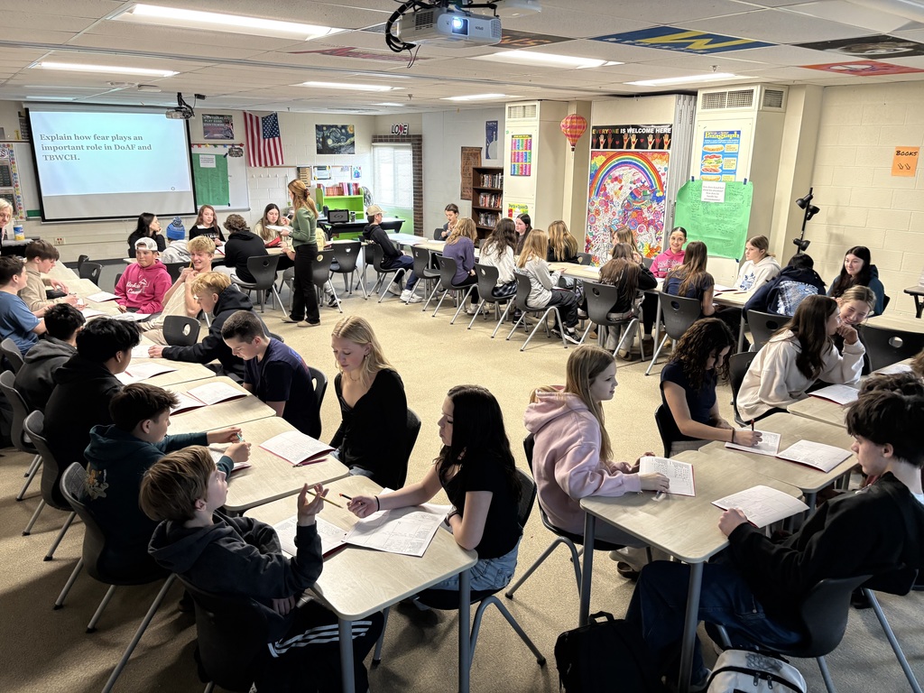 Students seated facing one another in desks to form a large rectangle in a classroom. Ms. Babiasz is standing toward the back of the rectangle