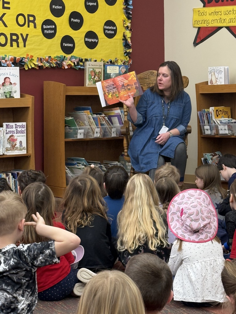 Author Miss Leestma reads her newly published book to a group of seated students in a library.