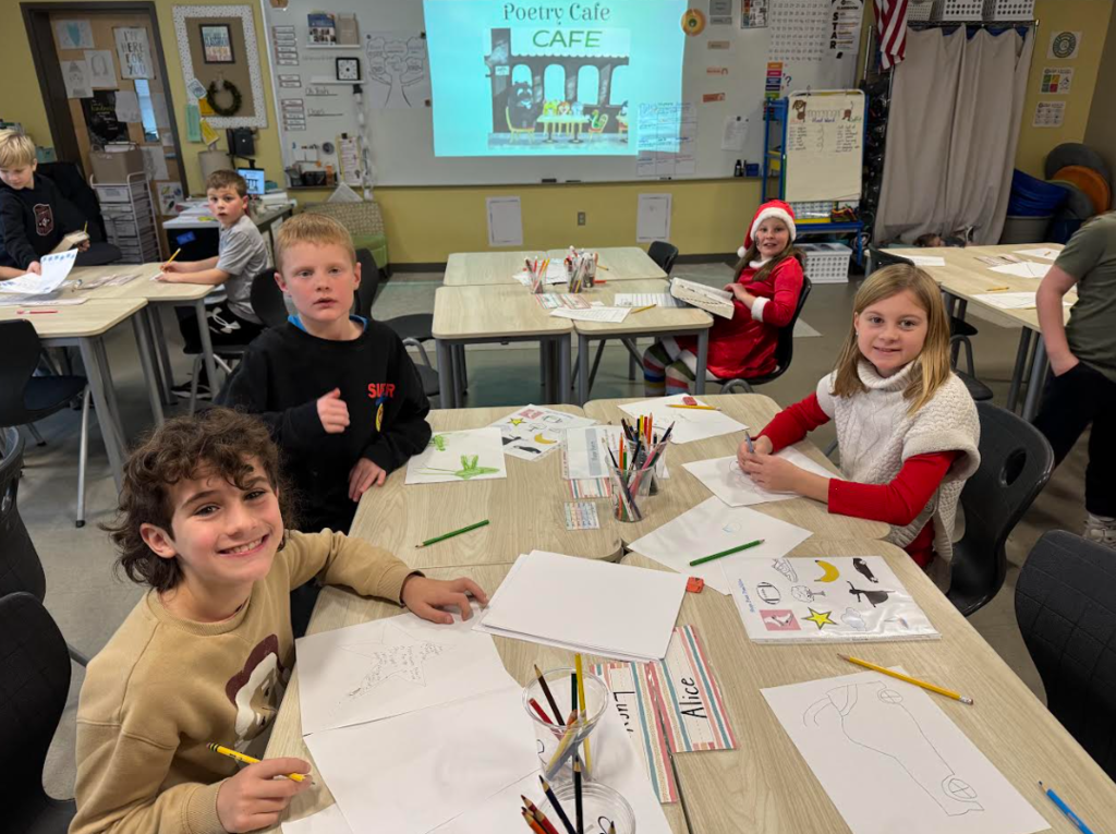 Students sit at light wood desks in a classroom "Poetry Cafe," working on illustrated poems with colored pencils. In the background, a projector displays a "Poetry Cafe CAFE" sign featuring a cartoon illustration of cats at a bistro. The students are smiling at the camera.