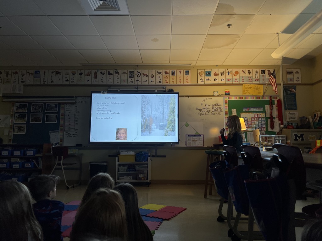  Gemini said A student stands in a dim classroom presenting a "Snow Day" poem on a digital screen to classmates seated on a colorful rug.