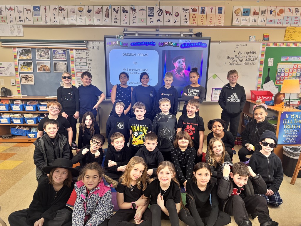 A 3rd-grade class dressed in black for a poetry slam. They pose in their classroom before a digital board listing poetic devices.