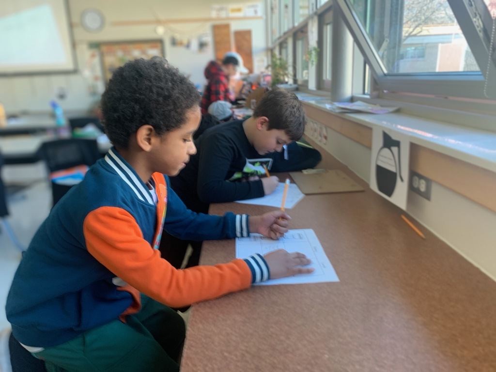 Two elementary-aged boys sit at a long wooden counter in a sunlit classroom, focused on writing on worksheets. The boy in the foreground wears a blue and orange varsity-style jacket and dark green pants, while the boy next to him wears a black long-sleeved shirt. Large windows to their right let in bright natural light, and other students are visible working in the blurred background.