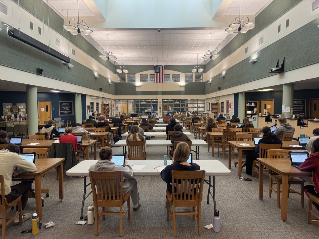 A wide-angle, high-angle shot of a spacious, well-lit school library or media center filled with students. Dozens of students are seated at long wooden and white folding tables, most with their backs to the camera, working on open laptops. The room features high ceilings with a large central skylight, green upper walls, and several circular hanging light fixtures. An American flag hangs prominently on the far wall above a set of glass doors and windows. The atmosphere is quiet and studious.