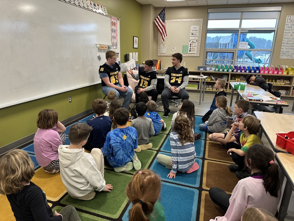 Three Traverse City Central football players are sitting in front of a class of second graders reading a book. The player in the middle of the three is holding a book out to his side reading while the other two players look on.