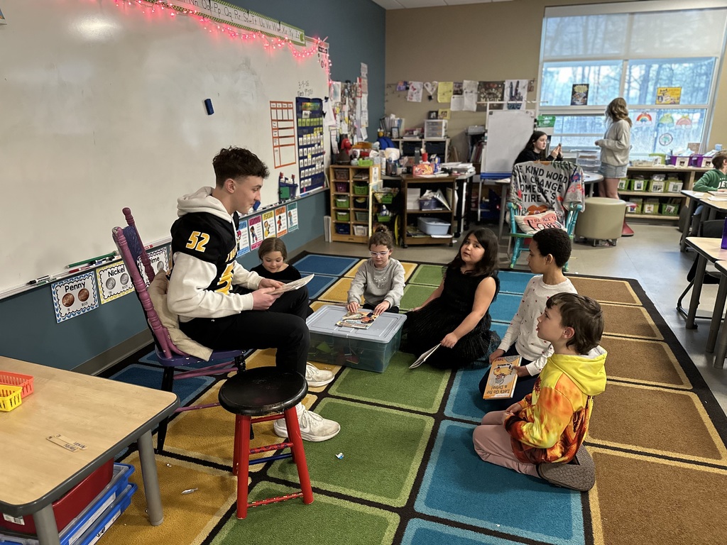 A Traverse City Central football players sits in a wooden chair holding a book in his lap while five students sit in front of him looking up at him and listening.