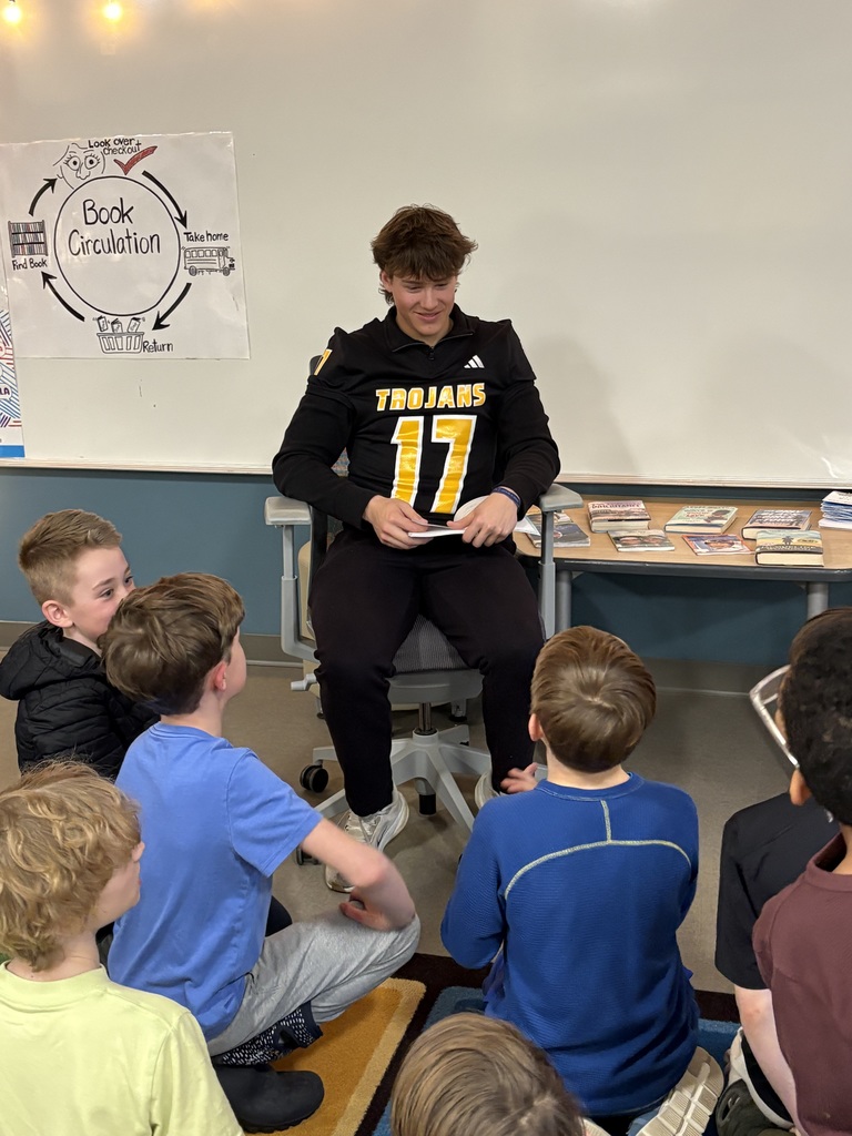 A Traverse City Central football player is seated in a chair holding an open book; he's seated in front of a small group of students who are looking up to him.