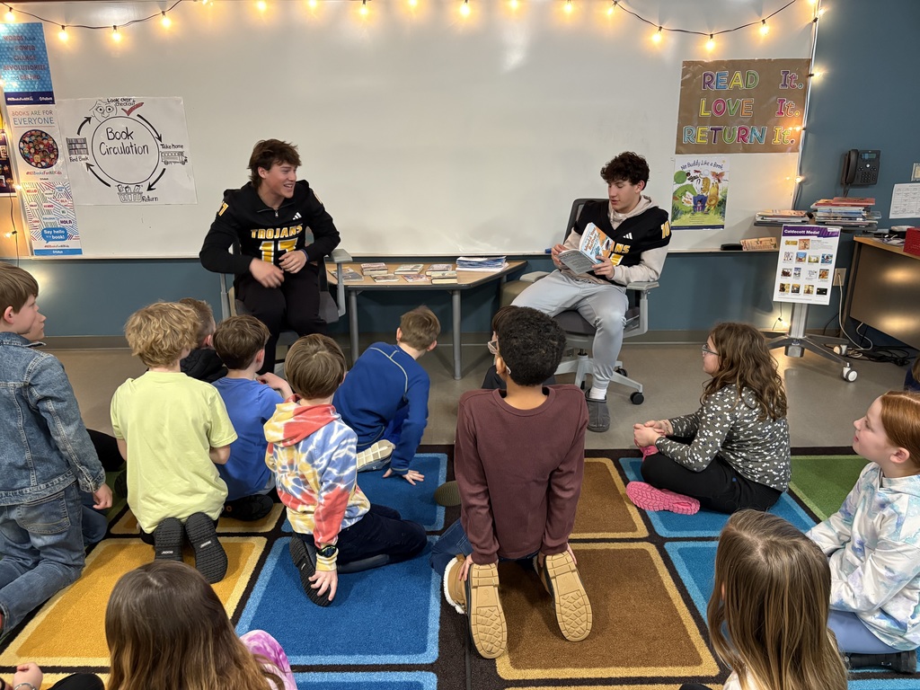 Two Traverse City Central football players sitting at the front of a classroom reading to a group of third graders who are sitting in front of them. The player on the right is reading from an open book while the player on the left looks on.