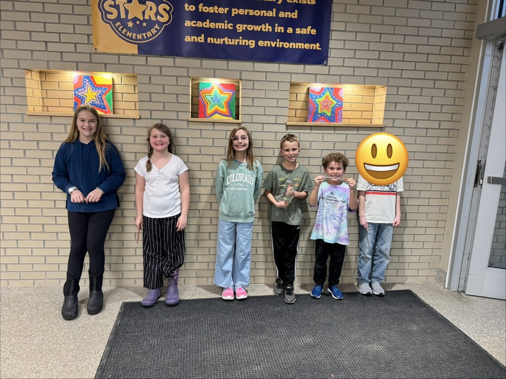 Six students standing against a brick wall, one each for grades kindergarten through fifth grade. The student second from the right is holding a star of the week pencil between his two hands. The student on the right, who is on a no-photo list, has a smiley face emoji sticker over his face.