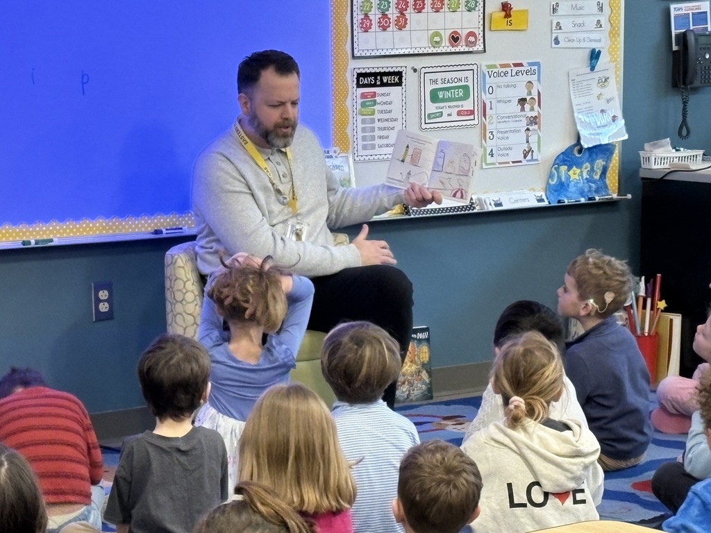 Mr. Swegles is sitting in front of Mrs. Edwardson's kindergarten class reading a book that he is holding out to the side.