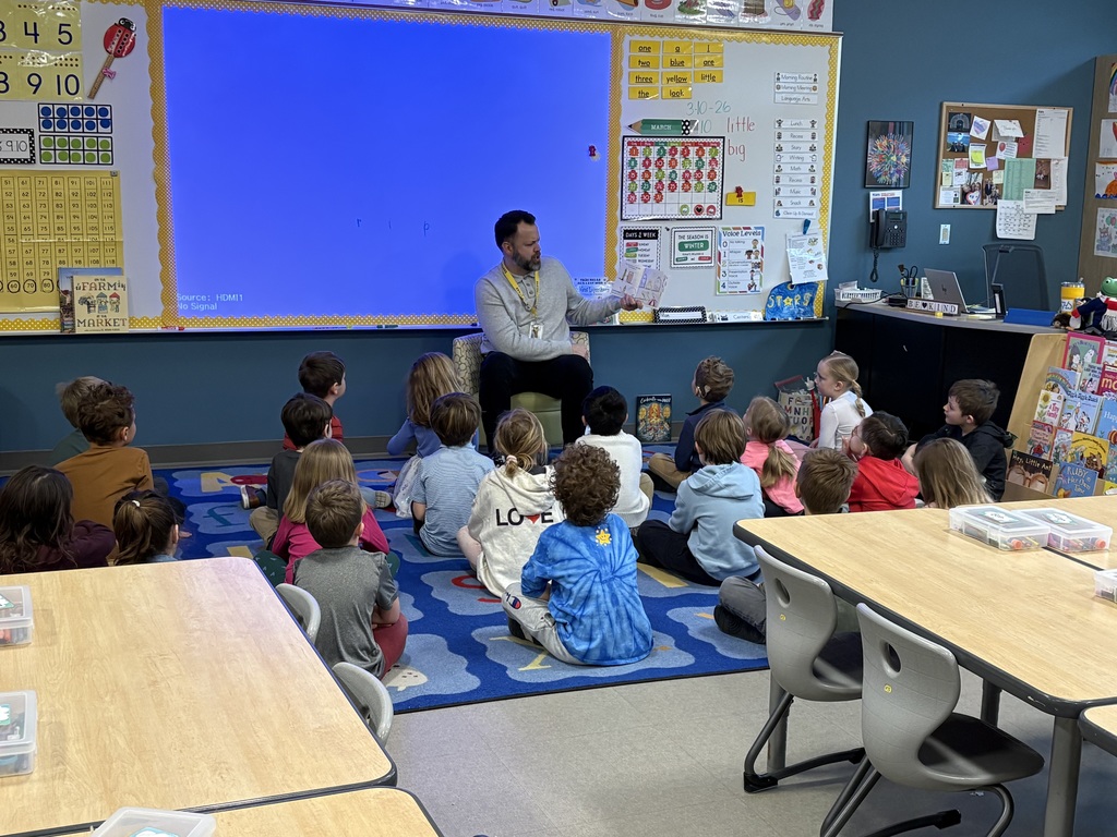 Mr. Swegles is seated in front of the class holding a book out to his side and reading to the students sitting on the rug in rows in front of him.