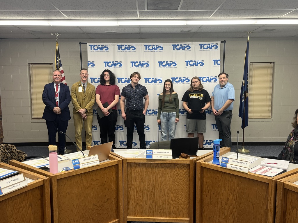 A group of seven people stands in front of a white TCAPS (Traverse City Area Public Schools) backdrop in a boardroom setting. The group includes four students and three staff members or officials.