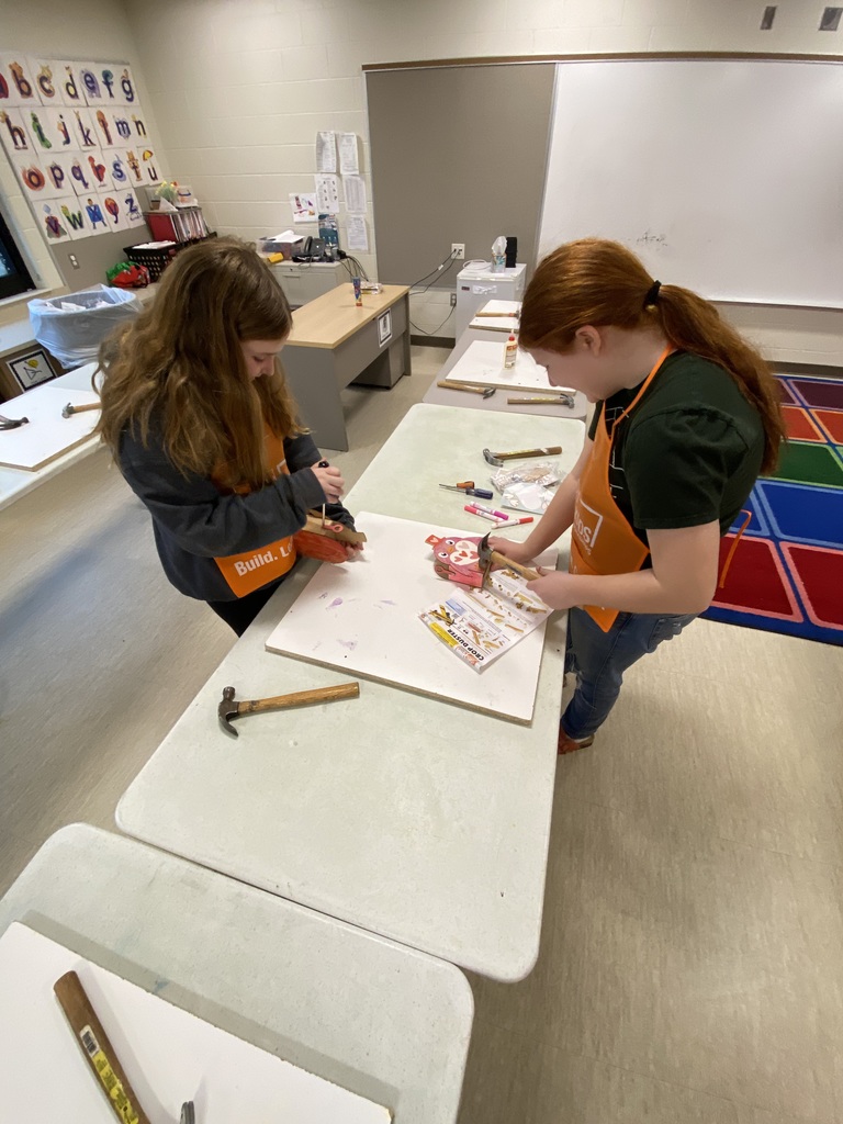 Two students working at a table with a hammer and instrucations.  They are weraring orange home depot smocks and building a pink owl box.  Behind them is a red, blue, green, orange and purple rug.