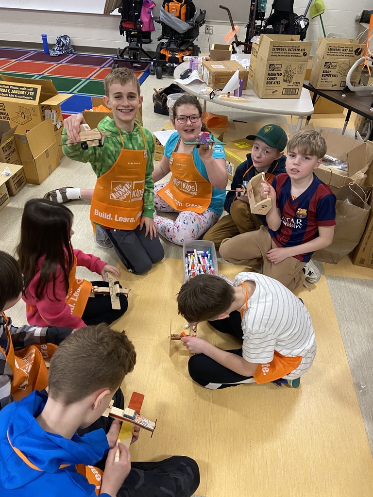 Studentst sitting on the floor of an empty classroom, filled with boxes and bags, creating a craft.  The students are wearing orange Home Depot  smocks and looking at the camera holding their wooden craft project that they made.  In the middle of the circle of student is a rubbermaid bin with markers.  One student is coloring on his wooden air plane craft.
