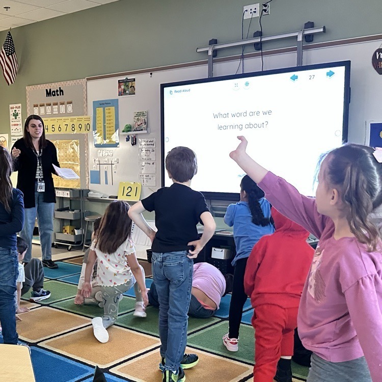 A teacher and her elementary students standing on the classroom rug as they listen to her teach.