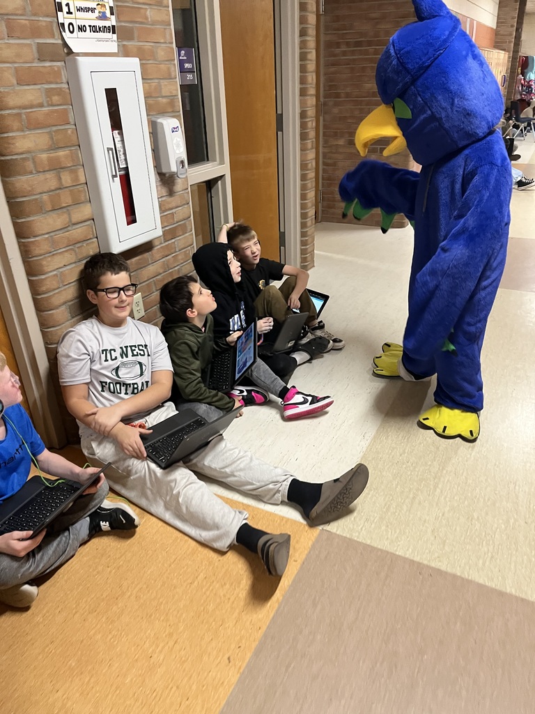 Stuents sitting in teh hallway reading books with their mascot standing by them