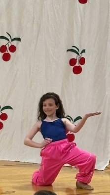 A young girl in a blue top and bright pink pants strikes a graceful dance pose on the stage.