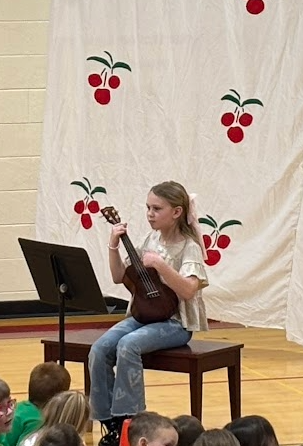 A girl with a pink hair bow sits on a bench while playing a ukulele for her performance.