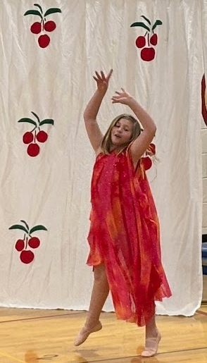 A girl in a flowing, colorful red and orange dress leaps mid-air during a dance routine.