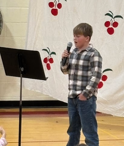 A boy in a grey plaid shirt sings into a microphone next to a music stand.