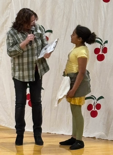 A woman in a plaid shirt with a clipboard introduces a girl in a yellow shirt to the audience.