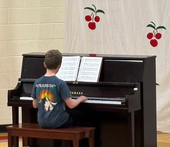 A boy in a "Steamboat" t-shirt playing a Yamaha piano from sheet music for an audience.