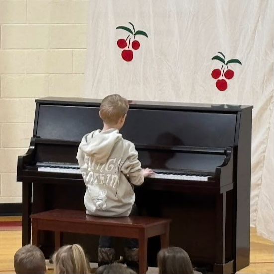 A student in a light-colored hoodie sits at a piano performing a solo for the crowd.