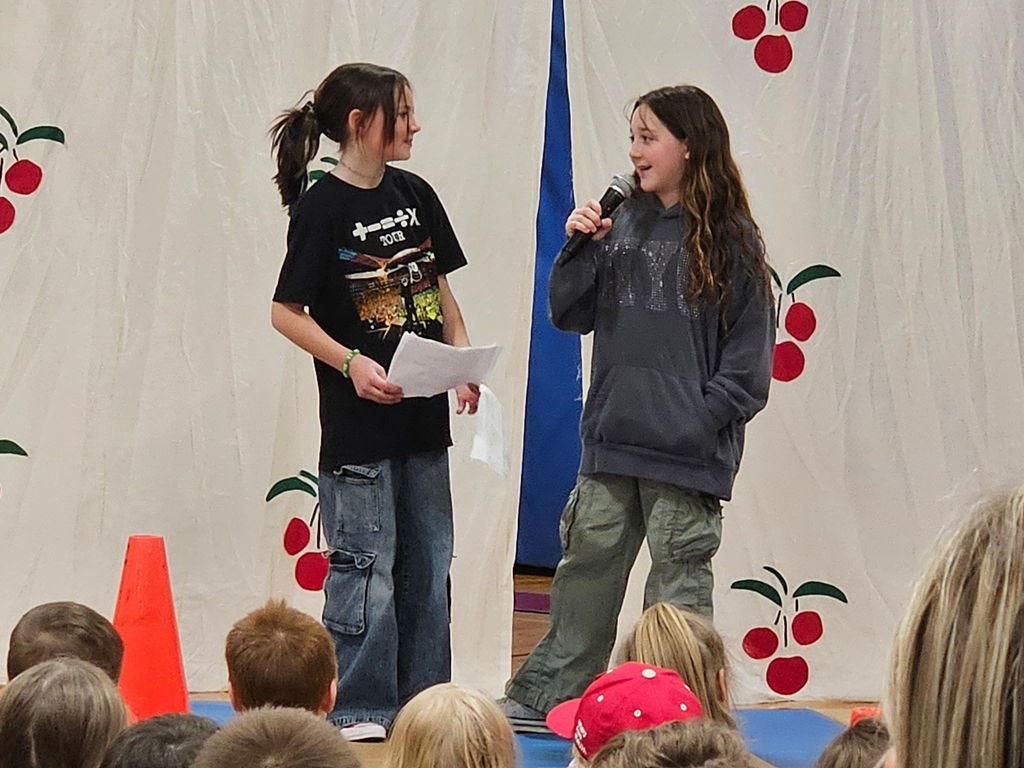 Two young girls smiling on stage, one holding a microphone and the other holding a script.