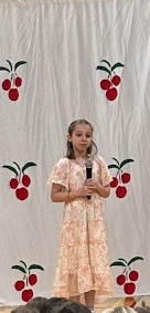 A girl in a floral peach dress stands on stage holding a microphone, ready to perform.