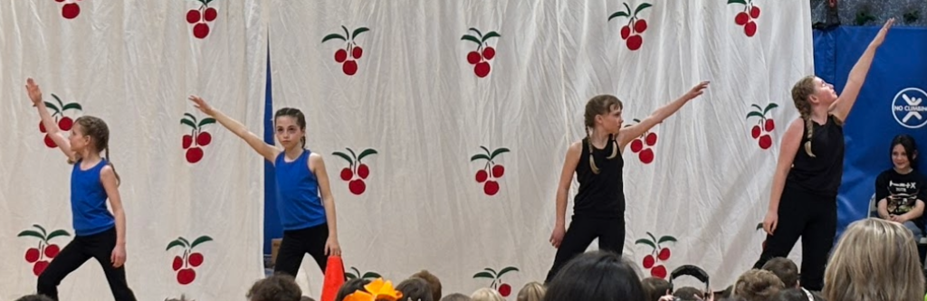 Four girls in blue and black dance attire strike a synchronized pose with arms raised on stage.