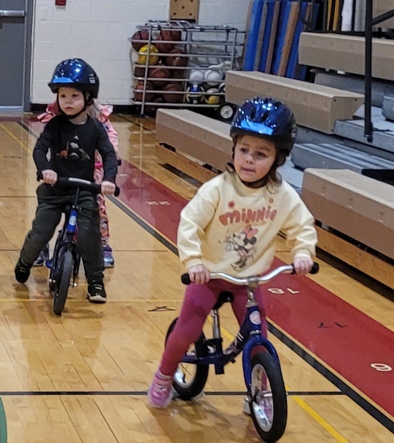 A girl in a Minnie Mouse sweatshirt leads a line of young bike riders across the school gym floor.