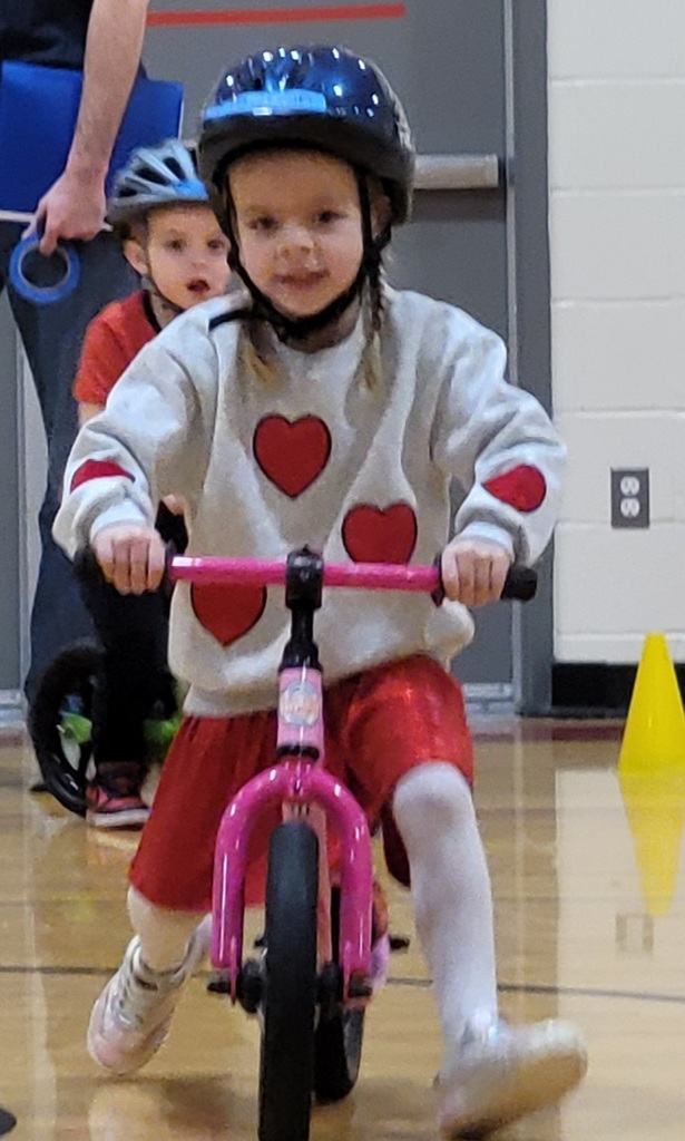 A young girl in a heart-patterned sweater successfully navigates a pink balance bike through the gym.