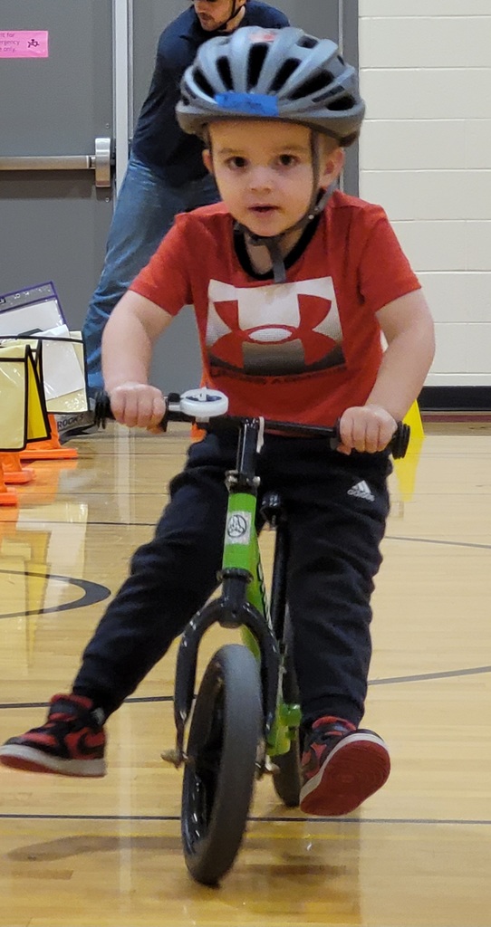 A preschooler in a red Under Armour shirt and grey helmet balances a green bike across the gym floor.