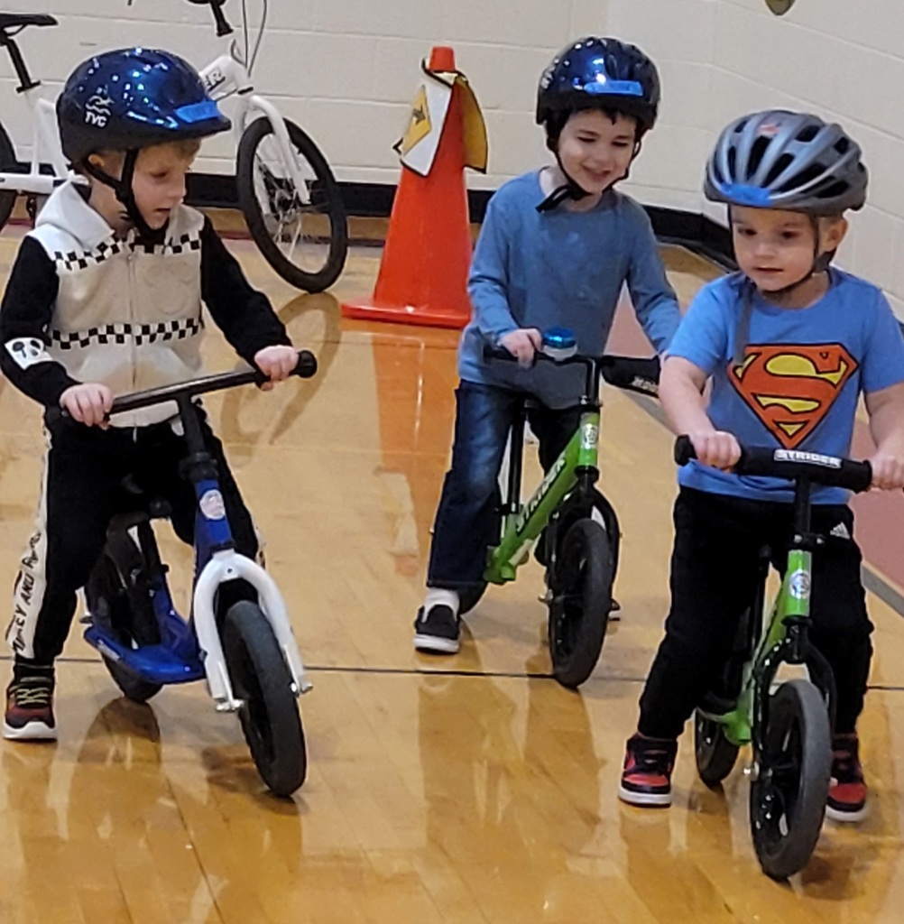 Three young boys in helmets and superhero shirts concentrate while riding their balance bikes in a row.