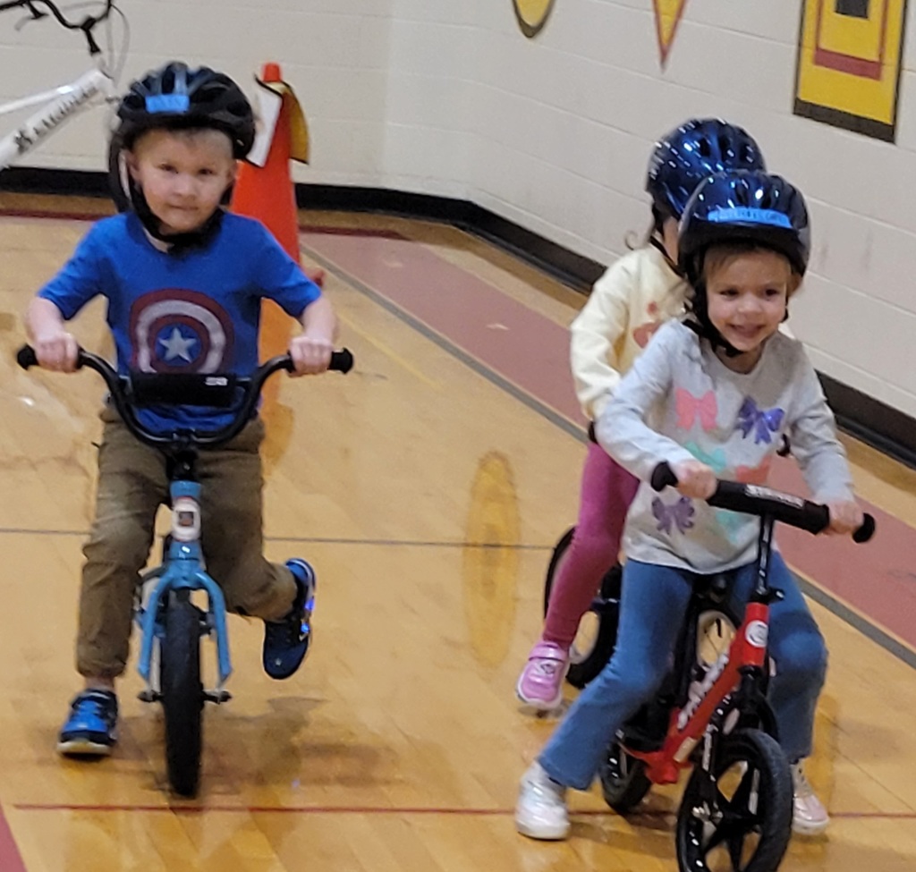 A boy in a Captain America shirt and a girl in a grey sweater race their bikes across the gym.