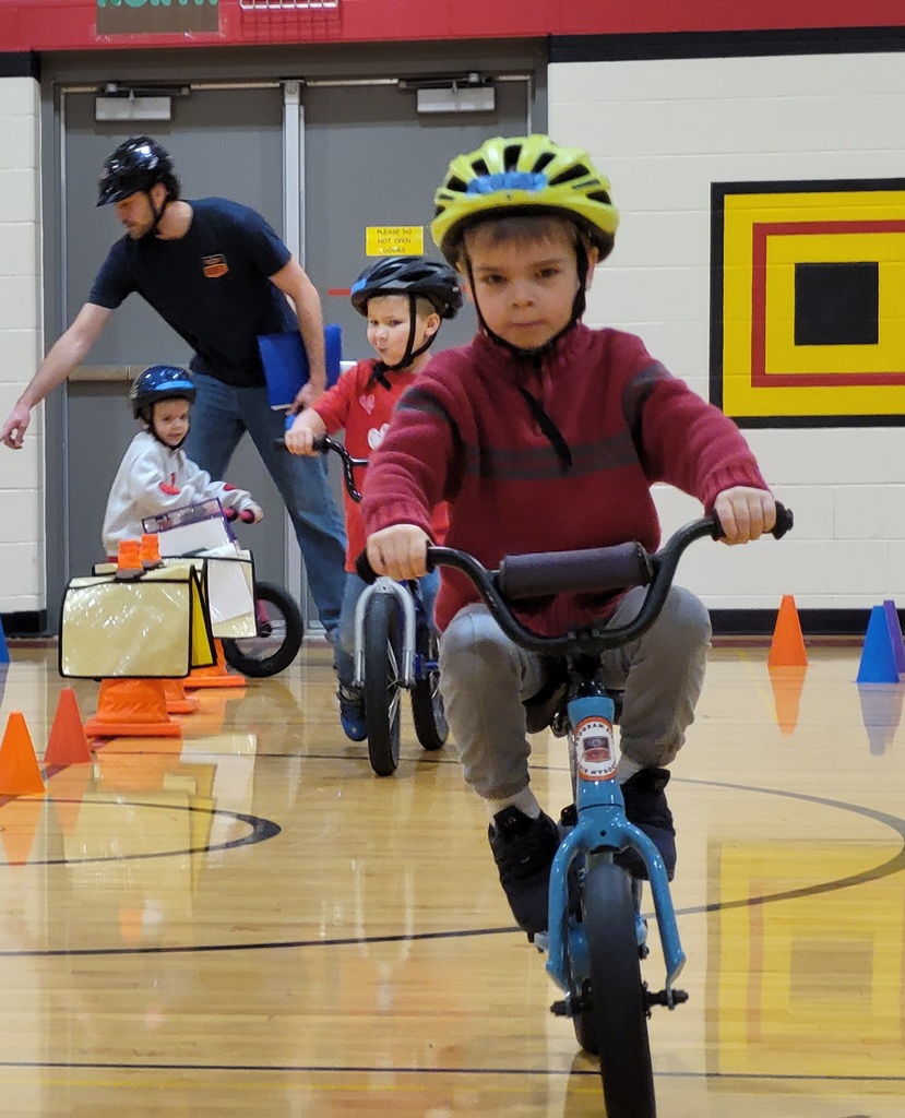 A line of children in helmets ride balance bikes past orange cones during their final gym practice.