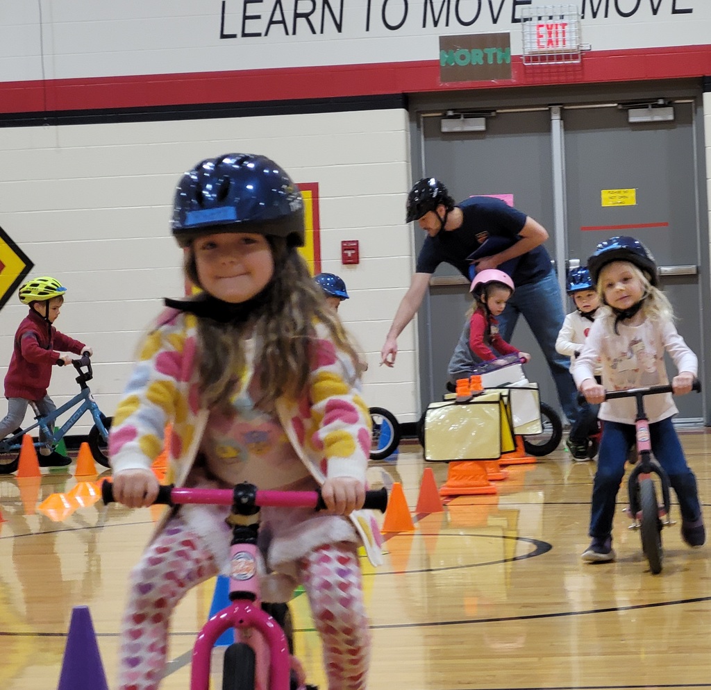 A girl in a heart-patterned sweater leads a group of preschoolers on bikes under a "Learn to Move" sign.