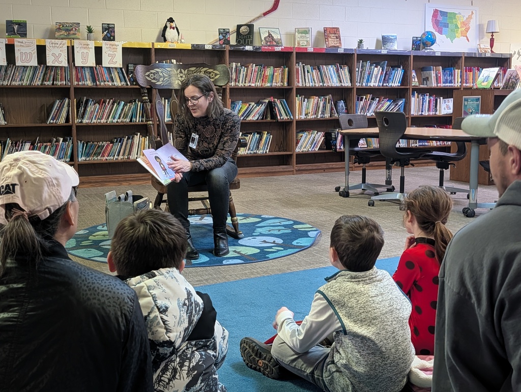 A woman in a patterned sweater reads a picture book to a group of children sitting on a blue rug in a library.