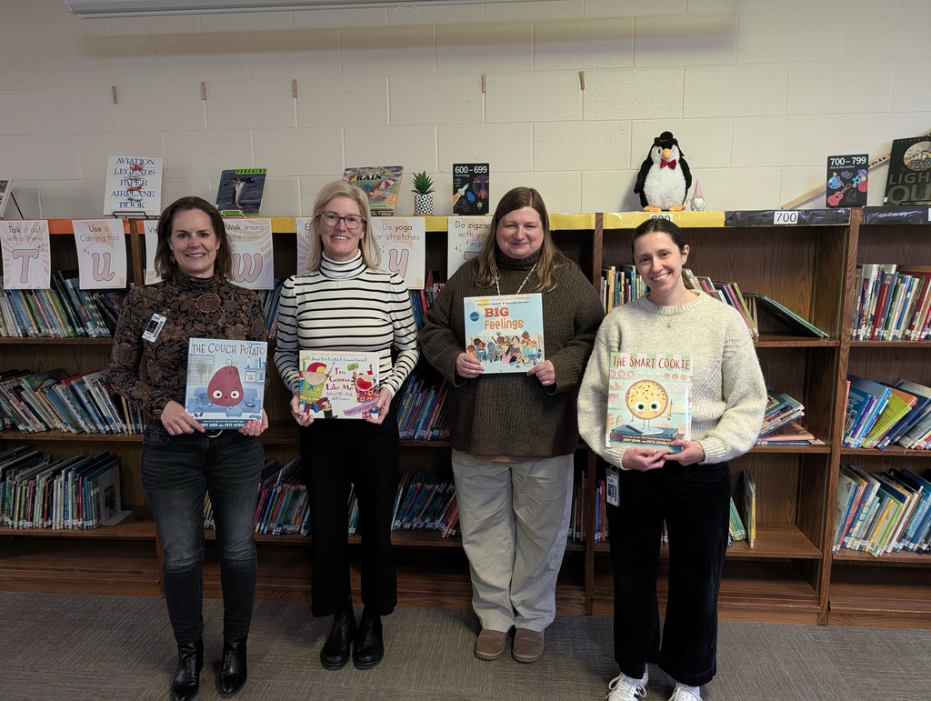 Four women stand in a library, each holding a different children's picture book in front of wooden bookshelves.