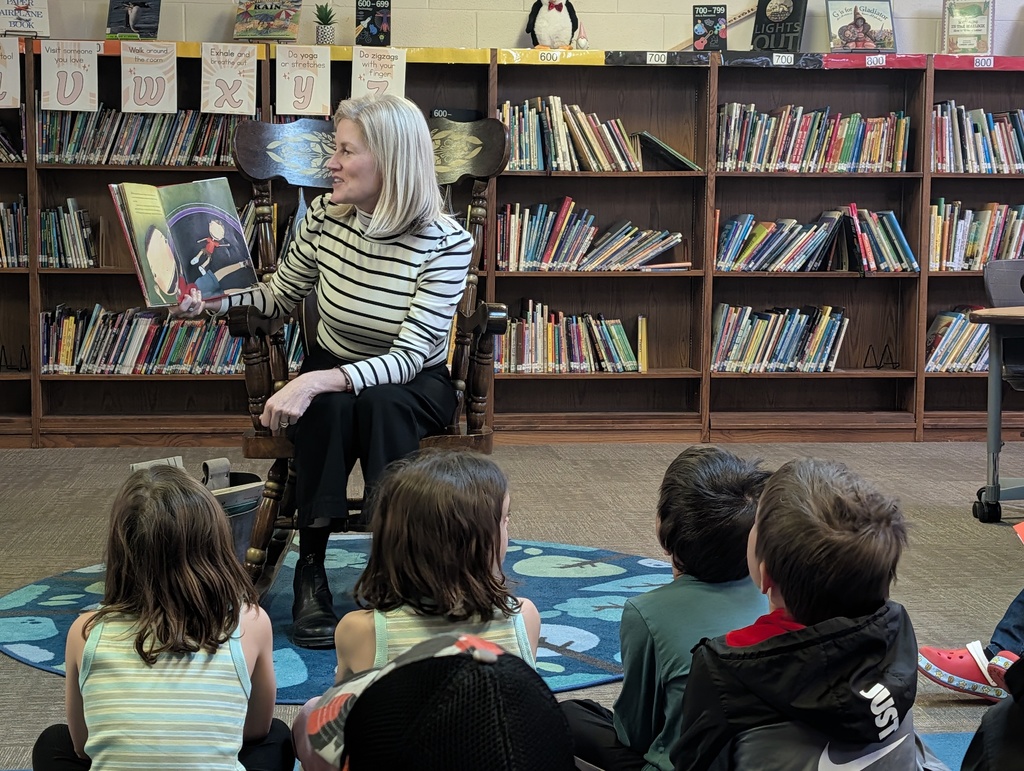 A woman in a striped shirt sits in a rocking chair reading a picture book to a group of children in a library.