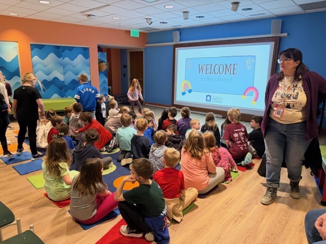 Students sitting on a colorful rug looking at a projection screen with the word welcome to the Traverse area district library.  A teacher is standing watching them.